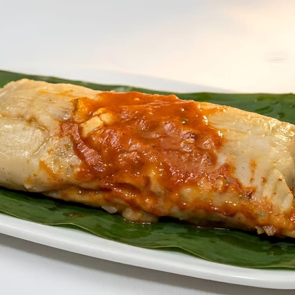 Tamale with red sauce on a banana leaf, served on a white plate.