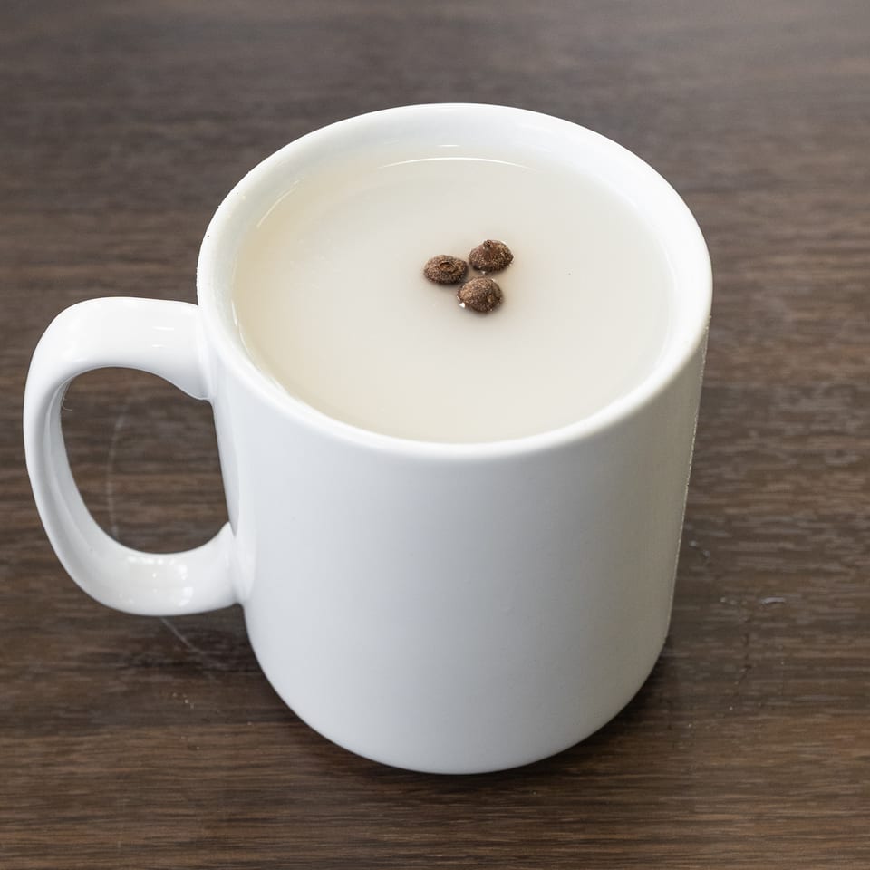 A white mug filled with a light-colored beverage, topped with three small brown seeds, on a wooden table.