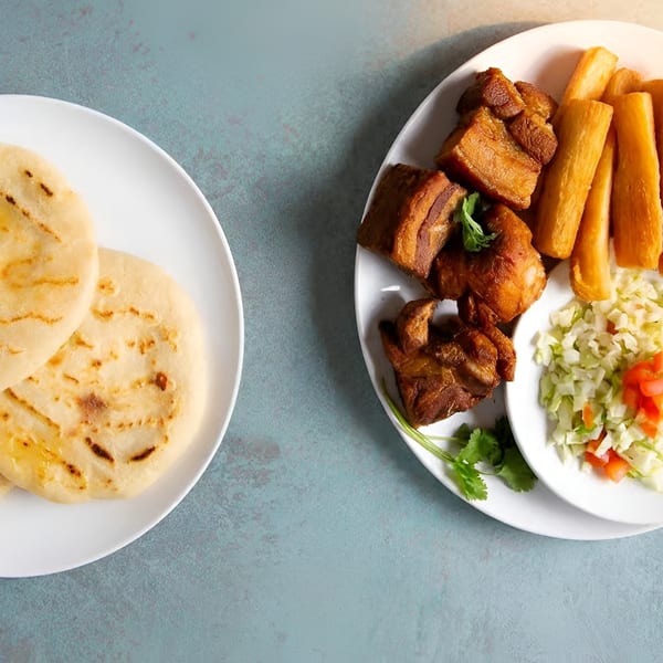 Plates of golden pupusas, fried pork, yuca fries, and curtido salad on a blue surface.