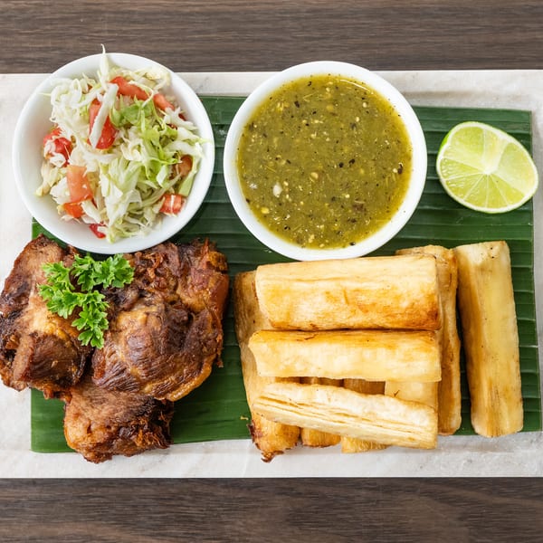 Fried pork, yuca fries, cabbage salad, and green sauce with a lime wedge on a banana leaf.