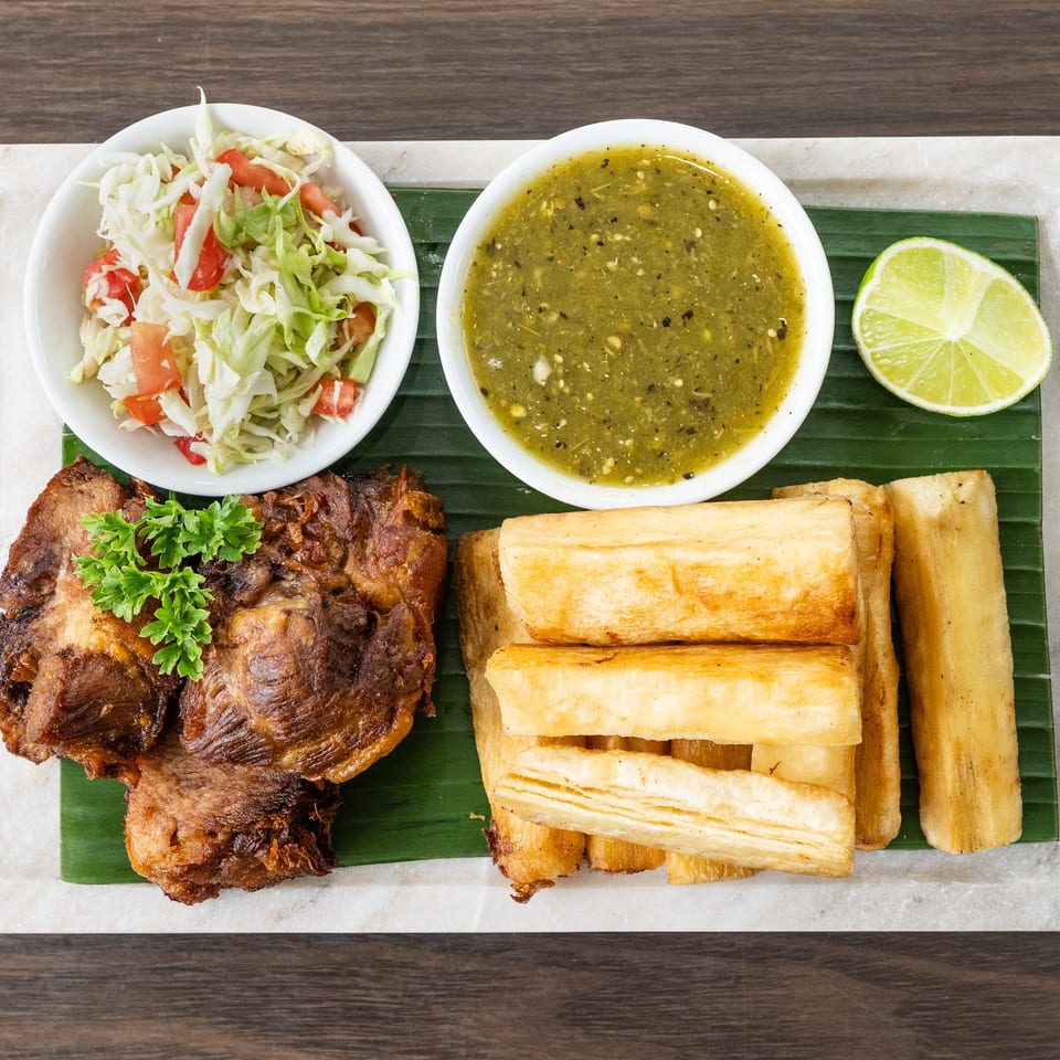 Fried pork, yuca fries, cabbage salad, and green sauce with a lime wedge on a banana leaf.