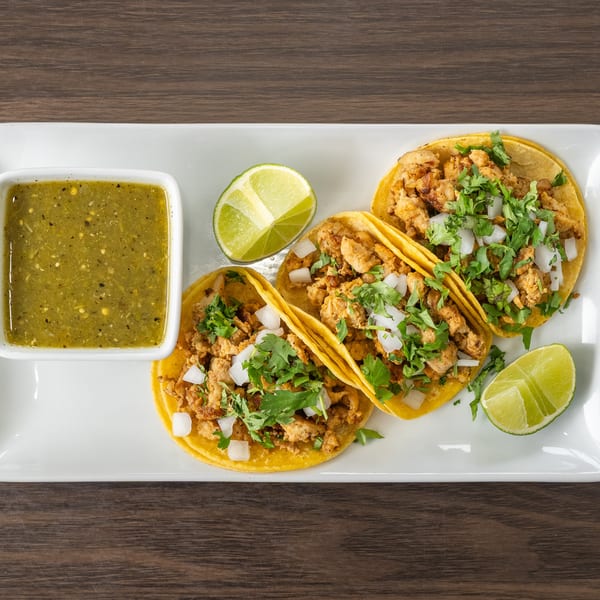 Three chicken tacos with cilantro, onions, lime wedges, and a side of green salsa on a white plate.