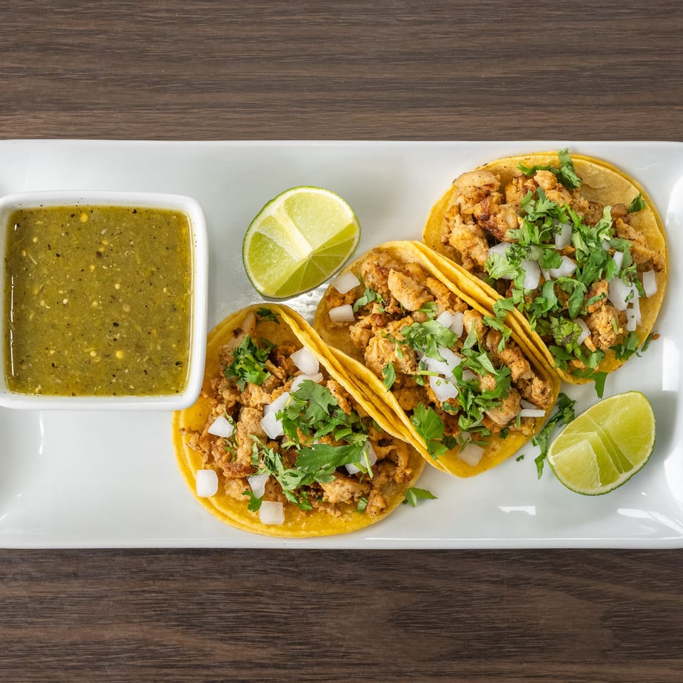 Three chicken tacos with cilantro, onions, lime wedges, and a side of green salsa on a white plate.